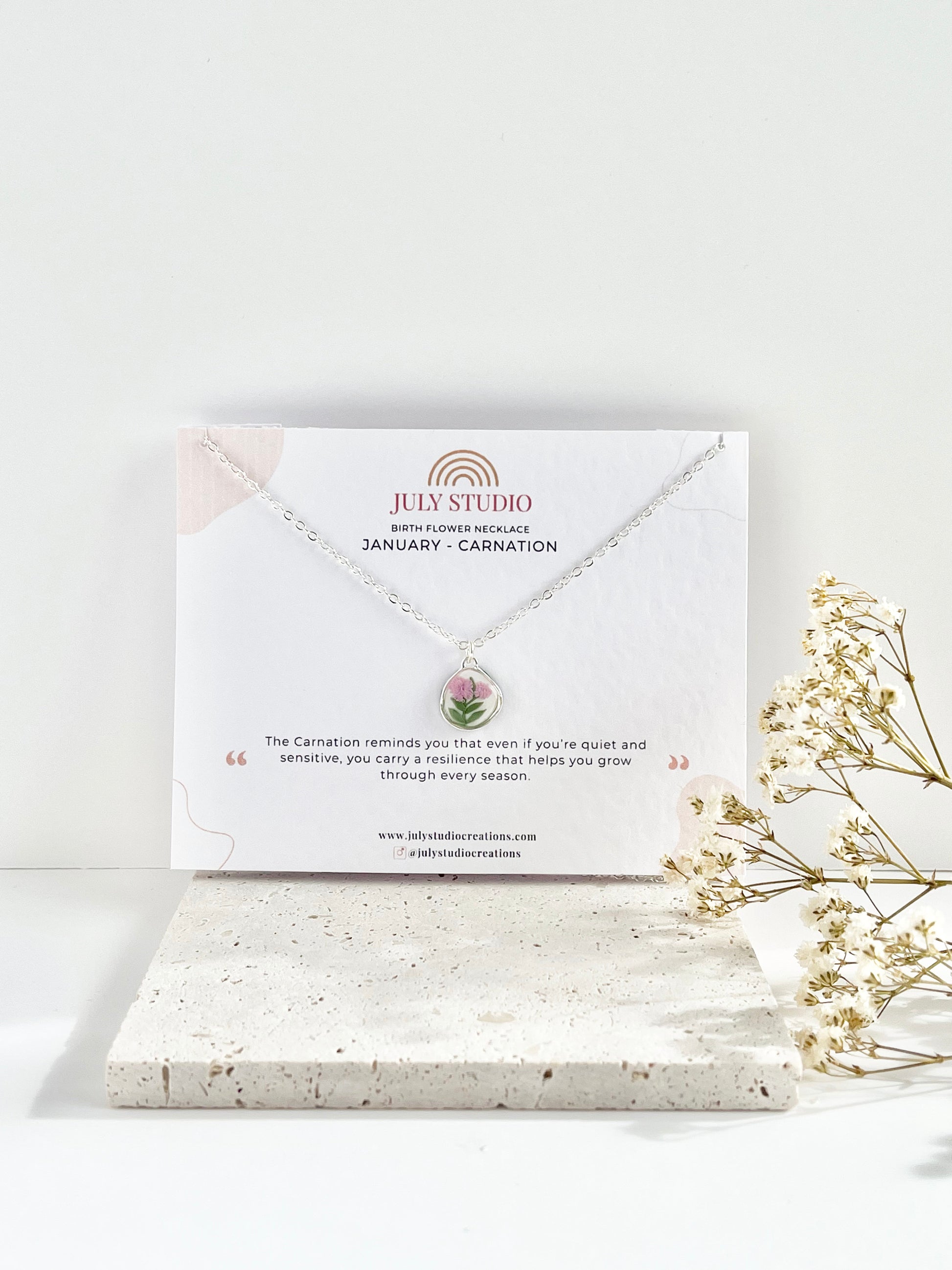 A silver clay necklace with a small carnation pendant on a jewellery card against a white background next to dried flowers